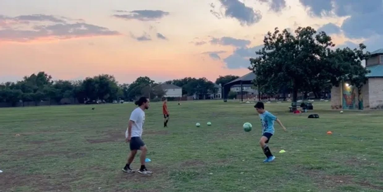 Children and an adult playing soccer on a grassy field at sunset.