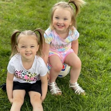 Two young girls with pig tails sitting in the grass. One of the girls is sitting on a soccer ball