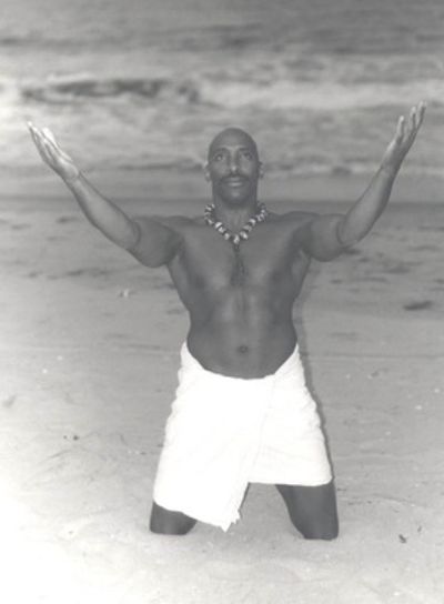 A man kneeling on the beach with arms raised, wearing a necklace and a cloth around his waist.