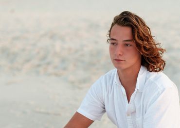 Young man with long hair wearing a white shirt, sitting on a sandy beach.