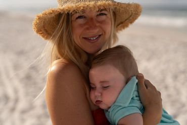 A woman wearing a hat holding a sleeping baby on the beach.