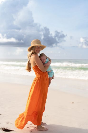 Woman in orange dress holding a baby on a sandy beach under a cloudy sky.