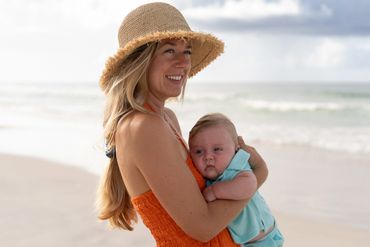 Woman in orange dress holding baby at the beach with a straw hat.