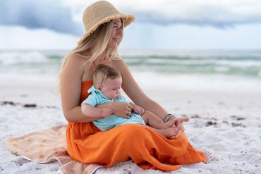 A woman in an orange dress holding a baby on a sandy beach.