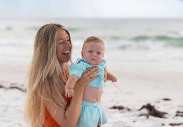 A joyful woman holds a baby in blue clothes on a beach.