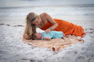 Mother lovingly interacts with her baby on a beach blanket by the ocean.