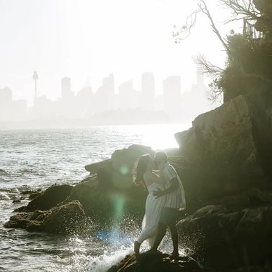 Couple sharing a romantic kiss on rocks by the water at sunset.