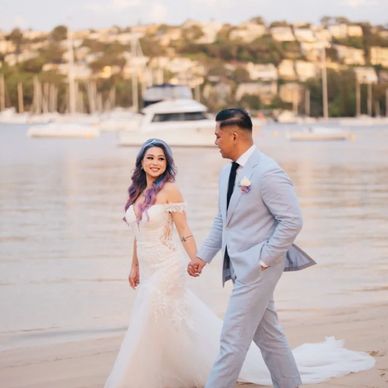 Bride and groom holding hands on a beach with yachts in the background.