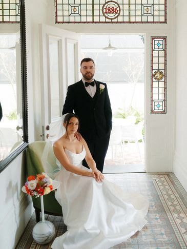 Bride in white gown seated beside groom in tuxedo in a bright room with stained glass.