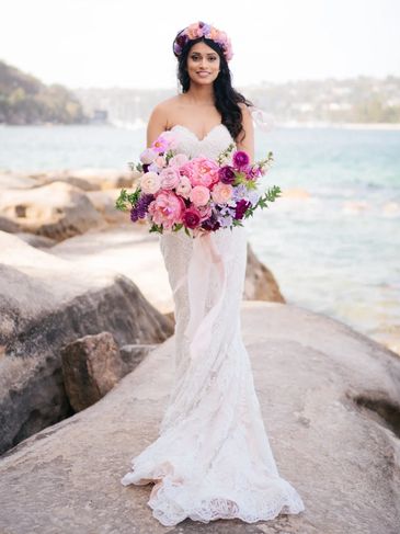 Bride in a white lace gown holding a vibrant bouquet by the seaside.