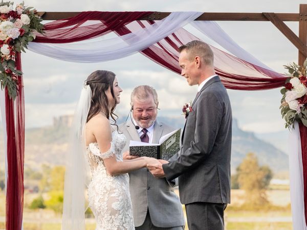 Bride and groom exchange vows under floral-decorated arch outdoors.