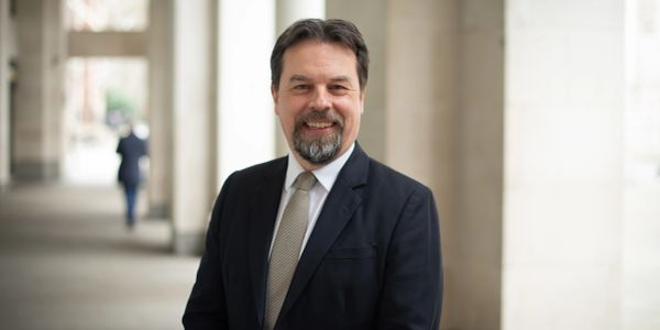 Smiling man in a suit standing outdoors near columns.