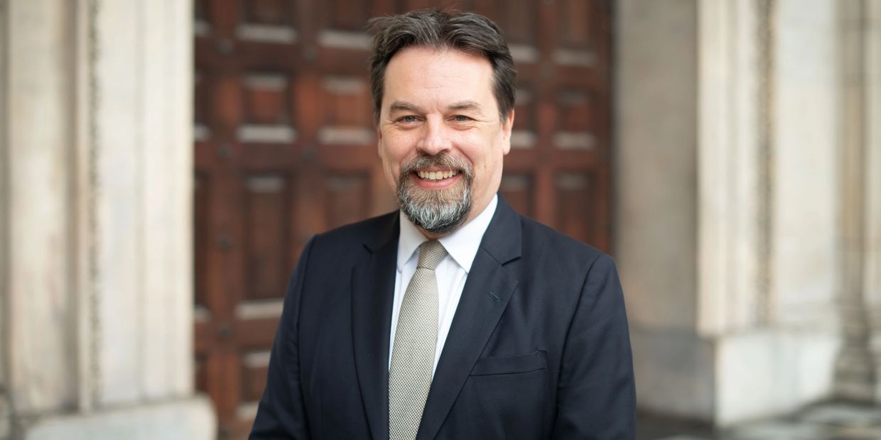 Smiling man in a suit and tie standing outdoors against a wooden door.