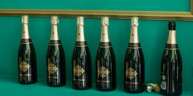 Sechs Champagnerflaschen in einer Reihe vor grüner Wand.