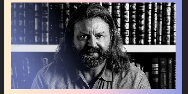 Black and white portrait of a bearded man with long hair in front of bookshelves.