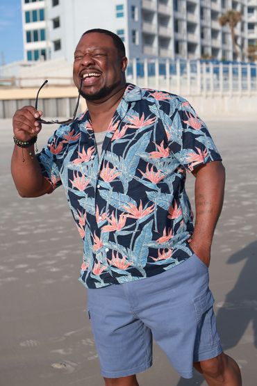 Man laughing on the beach in a floral shirt and blue shorts.