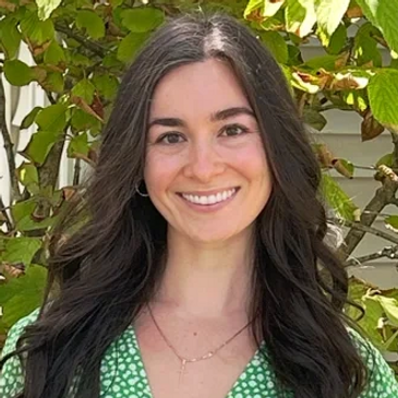 Smiling woman in a green floral dress stands outdoors with lush greenery behind her.