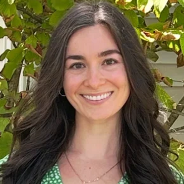 Smiling woman in a green floral dress stands outdoors with lush greenery behind her.