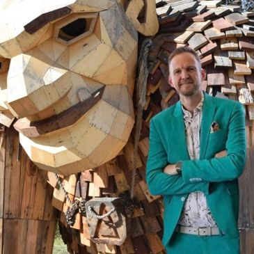 Man in teal suit standing next to a large wooden lion sculpture.