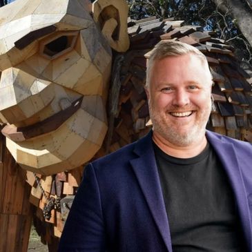 Smiling man in front of a large wooden sculpture of a face.