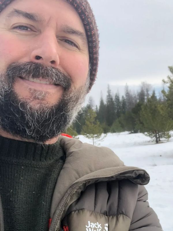 Bearded man in winter clothes smiling outdoors with snowy forest background.