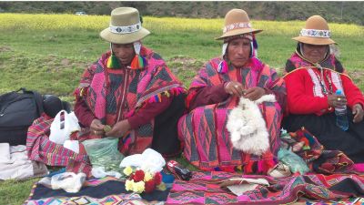 Three indigenous women in traditional clothing sitting and crafting outdoors.