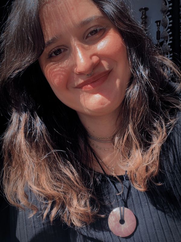 A woman with wavy hair and a septum piercing smiles softly in warm sunlight.