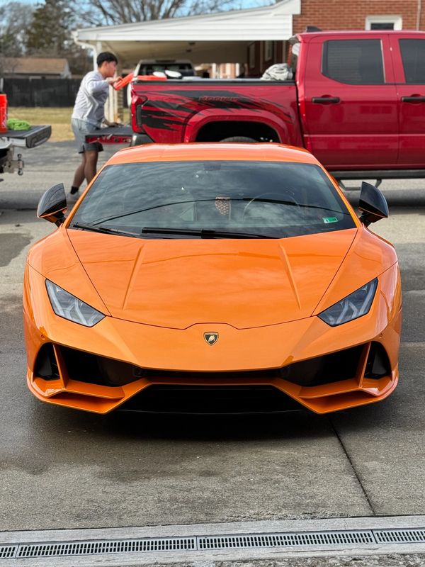 Bright orange Lamborghini parked in driveway with person and red truck in background.