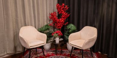 Two beige chairs with red flowers and green plants in between, set against dark and light curtains.