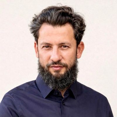 A man with a beard and curly hair wearing a dark shirt against a plain background.