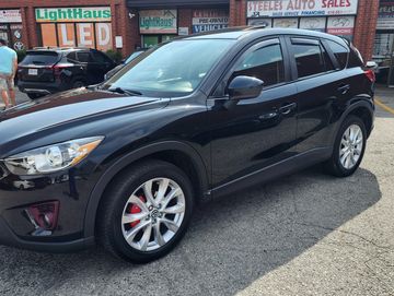 Black Mazda CX-5 SUV parked outside a car dealership with a For Sale sign.