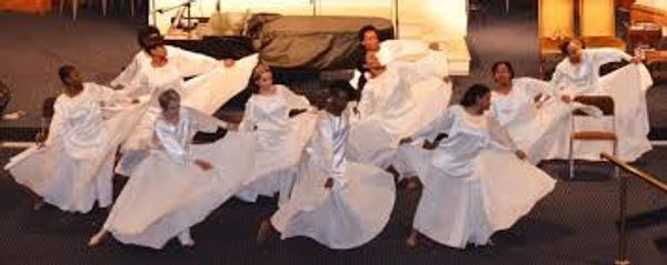 Group of dancers in white flowing dresses performing on stage.