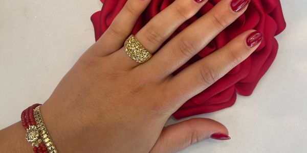 Hand with red nail polish, a gold ring, and bracelets on a red rose background.