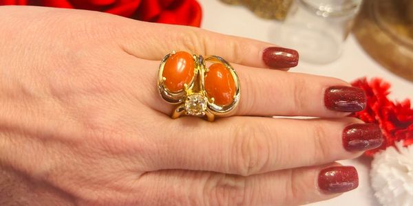 Hand with glossy maroon nails wearing a gold ring with two orange stones and a central clear gem.