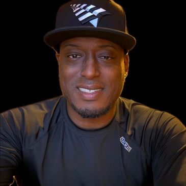 Man smiling, wearing a black cap and black shirt against a black background.