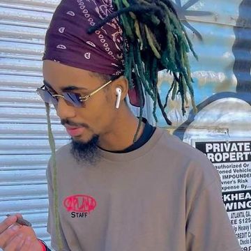 Man with green dreadlocks, sunglasses, and AirPods wearing a headscarf and casual shirt.