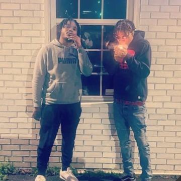 Two young men standing by a brick wall, one lighting a cigarette.