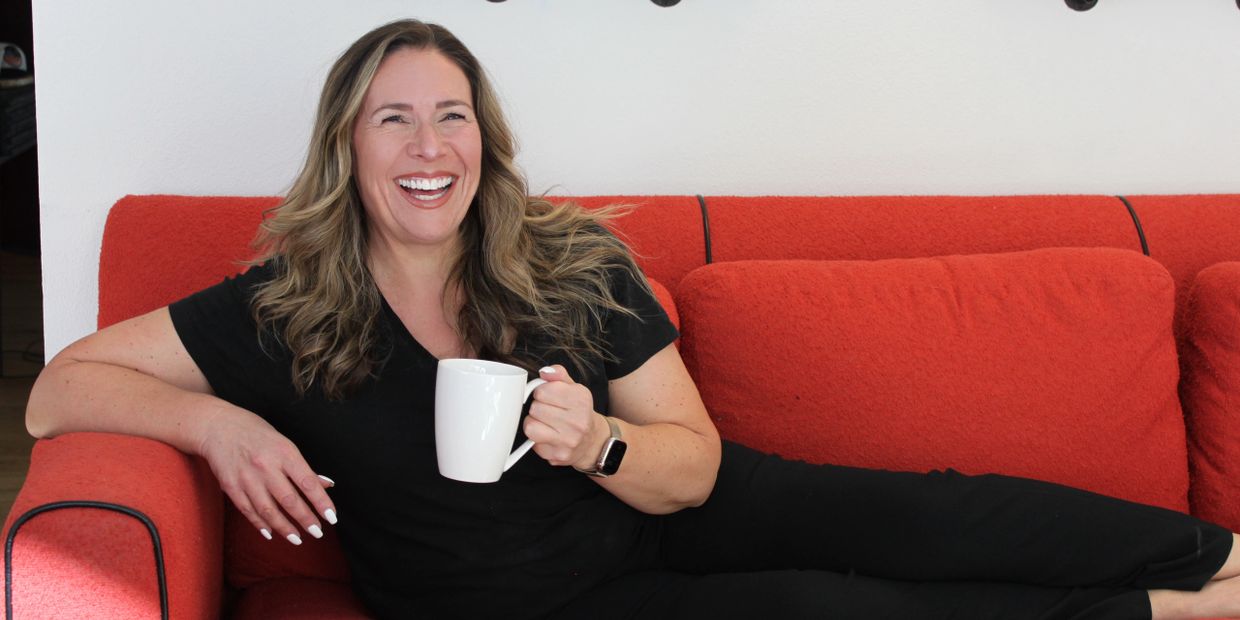 A woman in black lounging on a red couch, smiling and holding a white mug.