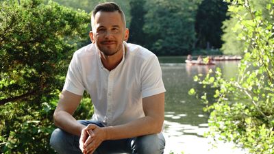 Man in white shirt sitting by a lake surrounded by greenery.