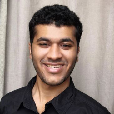 Smiling young man with short curly hair and a black shirt against a neutral curtain background.