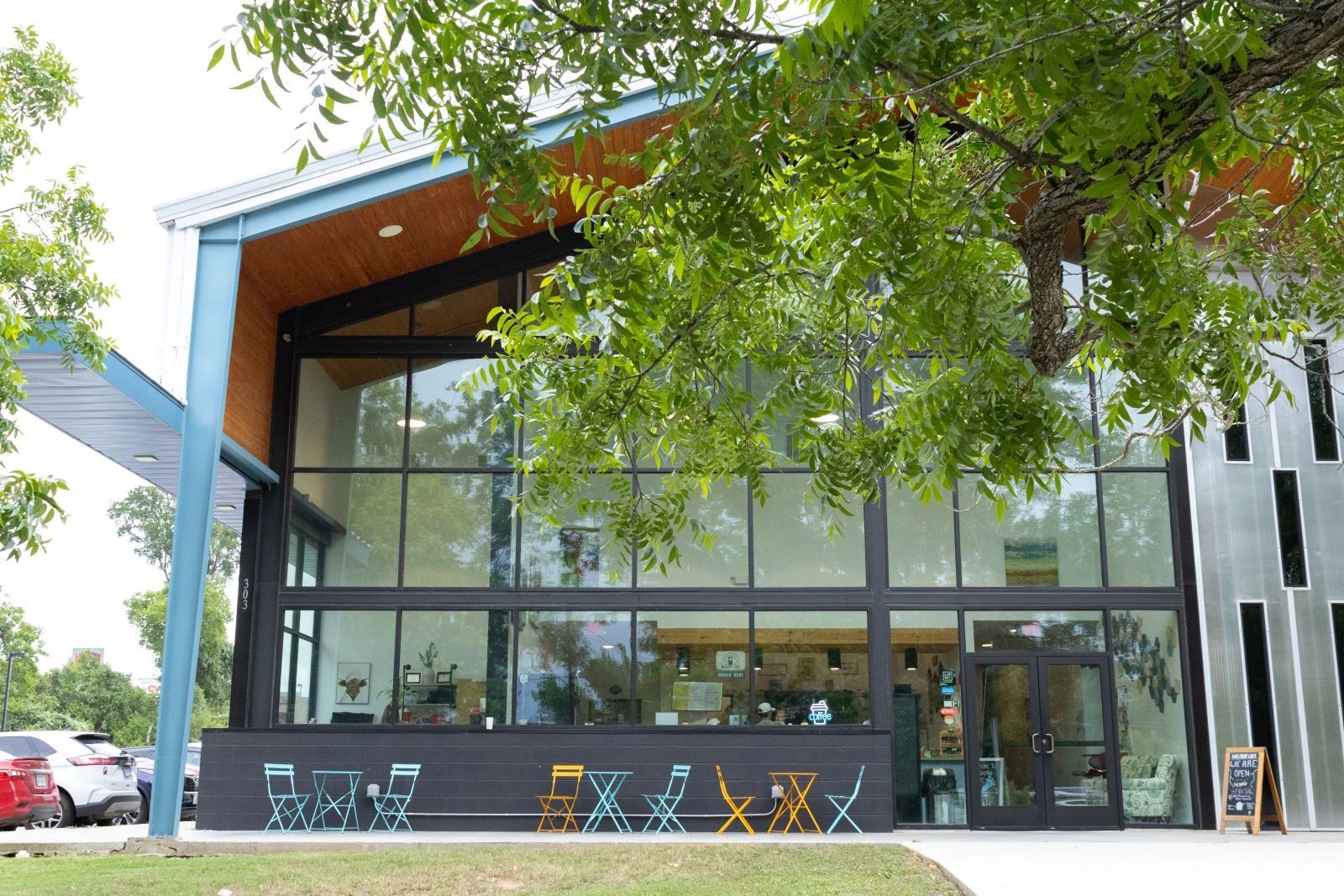 Modern coffee shop with colorful outdoor seating under green tree branches.