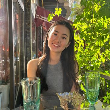 Young woman smiling at a sunlit outdoor café surrounded by green leaves.