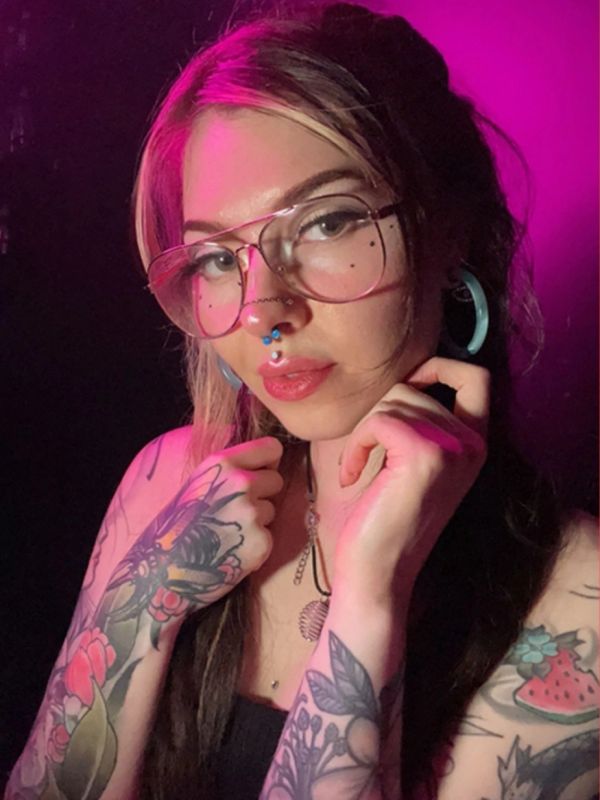 Woman with tattoos, glasses, and septum piercings under pink lighting.