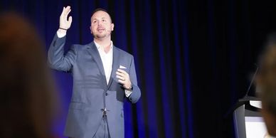 Man in suit giving a speech on stage with blue curtain background.