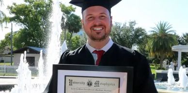 Graduate in cap and gown proudly holding diploma outdoors.