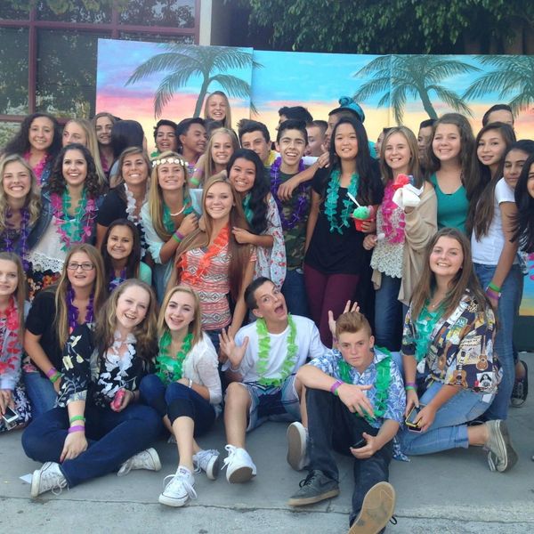 A large group of teenagers smiling at a tropical-themed party with leis and palm tree backdrop.