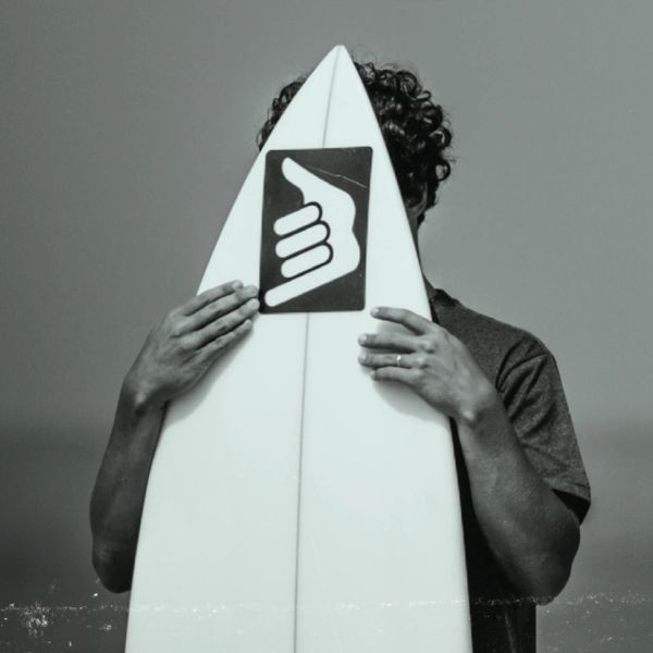 Person hiding face behind a surfboard with a hand symbol.
