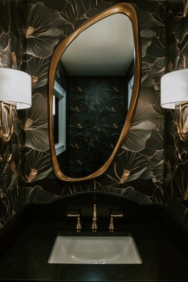 Powder room with floral wallpaper, organically shaped mirror, black counter, and modern wall sconces
