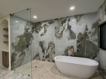 Freestanding tub in a glass‑enclosed walk‑in shower with green‑veined marble slabs; pebble flooring 