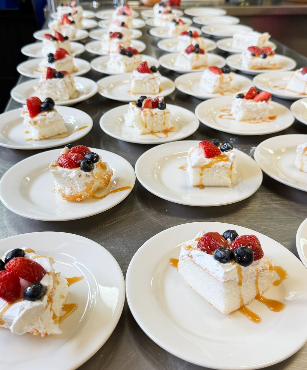 A table of beautifully-plated slices of angel food cake with whipped topping, fresh berries & syrup.